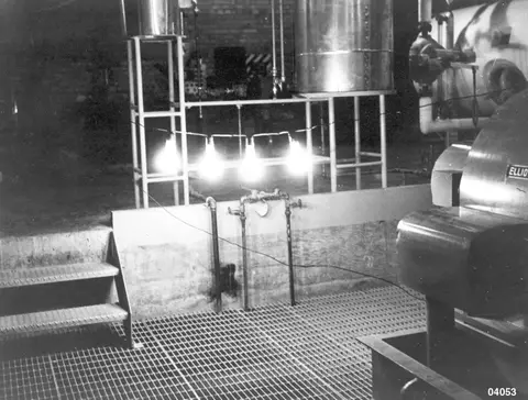 Black and white photograph showing the interior of an industrial room with metal grating flooring and machinery.