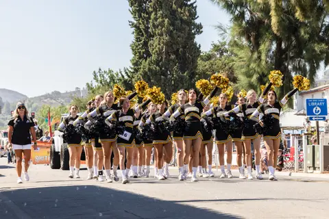 A cheerleading squad in black and gold uniforms marches in a street parade.
