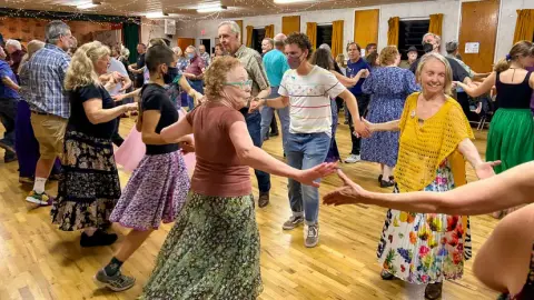 A group of people participate in folk dancing inside a hall with wooden floors.
