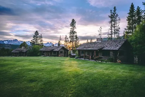 A photo of rustic log cabins situated on a large grassy lawn with mountains in the background.