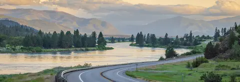 A scenic landscape view featuring a winding road next to a river and mountains under a cloudy sky.