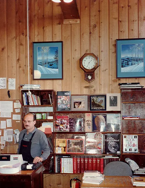 A man stands behind a counter in a room with wood-paneled walls and bookshelves.