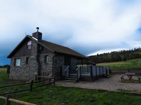 A stone cabin with a wooden deck stands in a grassy landscape under a cloudy sky.