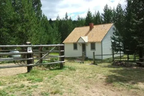 A small cabin stands in a clearing surrounded by trees and a wooden fence.