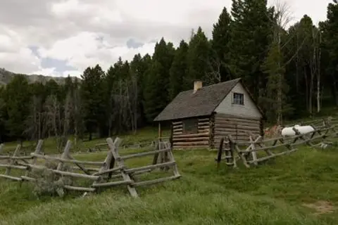 A rustic log cabin sits in a grassy meadow surrounded by a wooden fence and pine trees.