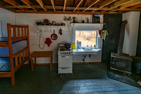 An interior view of a rustic cabin with a bunk bed, small kitchen area, and wood-burning stove.