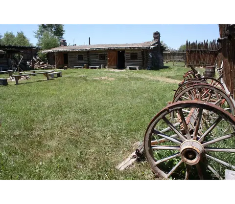 An old wooden cabin sits across a grassy yard with wagon wheels in the foreground.