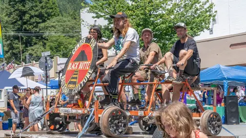 People ride a custom multi-person bicycle contraption on a street during an outdoor event.