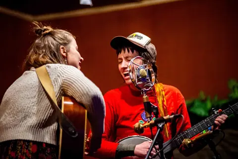 Two musicians perform on stage together, singing into a vintage microphone while playing guitars.