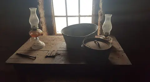 An interior view showing a wooden table with lamps, a basin, and a pot in front of a window.