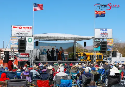 A band performs on an outdoor stage before a seated crowd beneath an American flag and an Arizona flag.