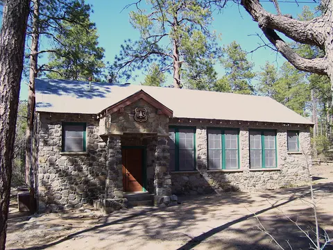 A stone building with a metal roof and green-trimmed windows stands in a forest clearing.