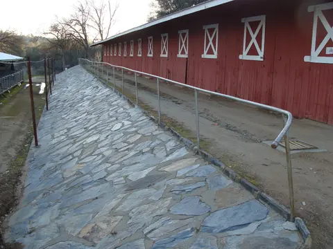 An exterior view of a long red barn with a stone embankment and a metal handrail.