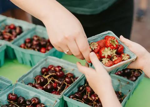 A person handing a basket of strawberries to another person over a table filled with fruit baskets.