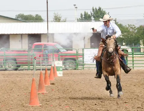 A cowboy rides a horse while firing a gun during an equestrian event.