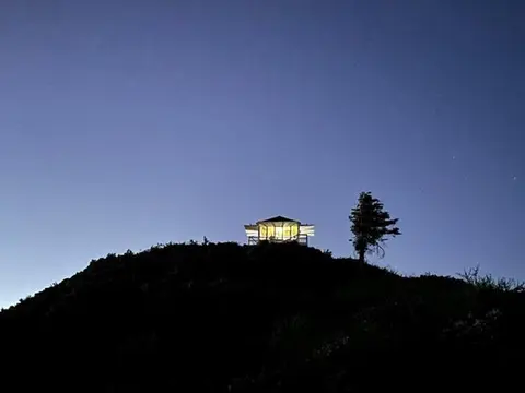 A brightly lit small structure stands atop a dark hill under a blue evening sky next to a single tree.