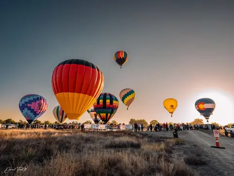 Several colorful hot air balloons launch from a field at sunrise as a crowd watches.