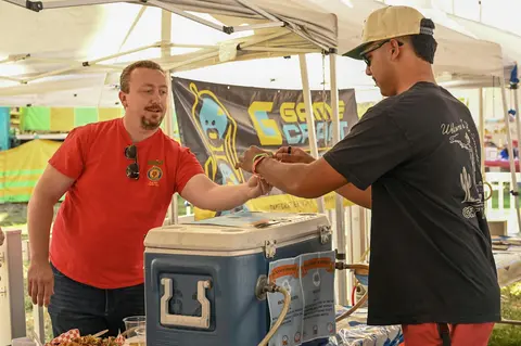 A man in a red shirt stands across from another man wearing a baseball cap at a outdoor booth.