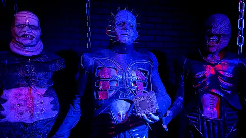 Three costumed horror characters stand in front of a brick wall under blue light.