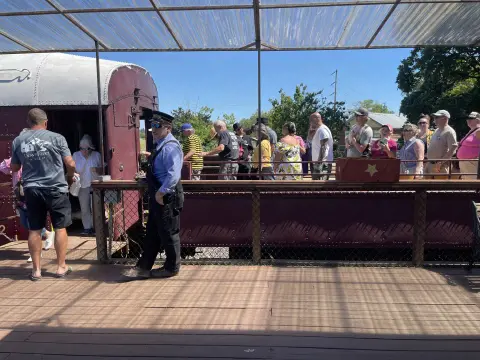 A conductor stands on a wooden platform next to passengers boarding a train car.