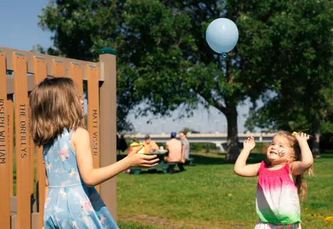 Two young girls play outdoors with a blue balloon near a park structure.