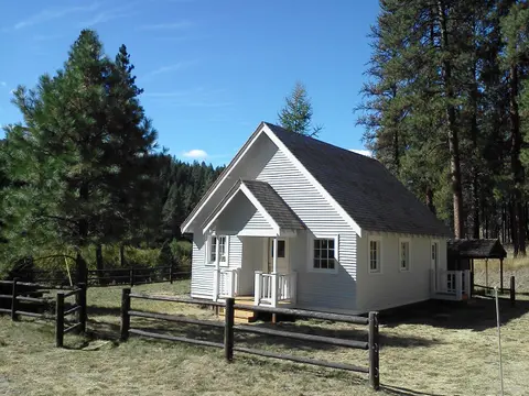 A small white building surrounded by pine trees and a wooden fence.