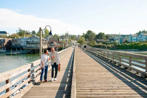 Two men walk along a wooden pier beside a waterfront town.