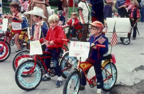 Children ride decorated bicycles in a parade holding American flags and signs.