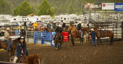 People and horses gather in a rodeo arena during rain.