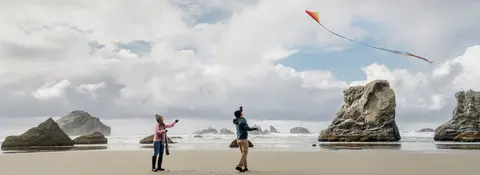 Two people fly a kite on a sandy beach with rock formations and ocean waves in the background.