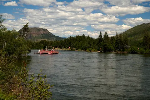A ferry crosses a wide river surrounded by trees and hills under a cloudy sky.