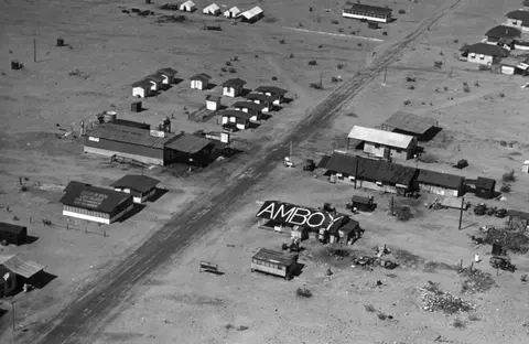 An aerial black and white photograph showing a small desert settlement with a large sign spelling AMBOY on a roof.