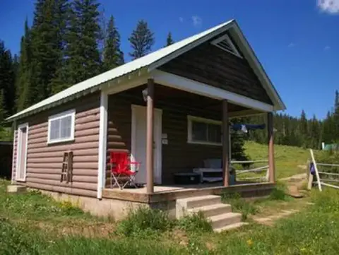 A log cabin with a covered front porch in a mountainous, grassy setting.