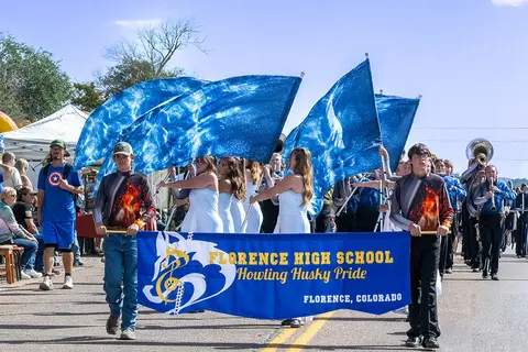 A high school marching band marches in a street parade carrying flags and a banner.
