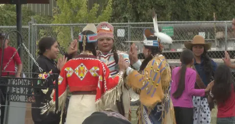 People in traditional attire and casual clothing gather outdoors near a chain-link fence.