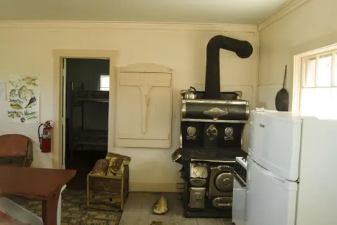 A rustic interior showing a vintage wood-burning stove, refrigerator, and a doorway leading to a bedroom with bunk beds.