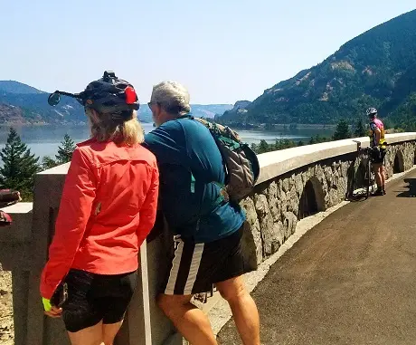 Two cyclists look out over a scenic body of water from a stone bridge.