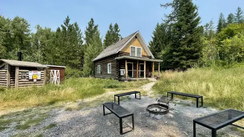 A rustic log cabin sits in a clearing surrounded by trees, with a fire pit and benches in the foreground.