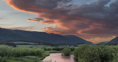 A river flows through a grassy valley beneath mountains and a cloudy sunset sky.