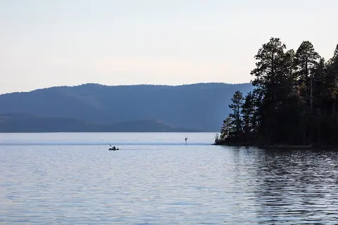 A person kayaking on a calm lake with forested hills in the background.
