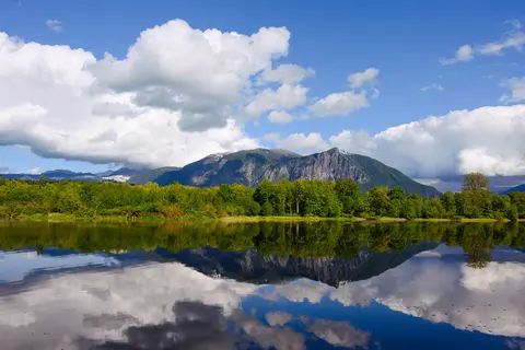 A scenic view of mountains and trees reflected in a calm body of water under a blue sky with white clouds.