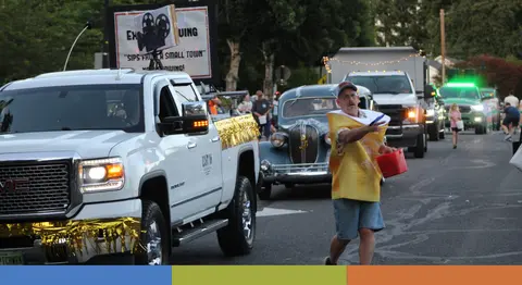 A person in a parade tosses items from a bucket near vehicles.