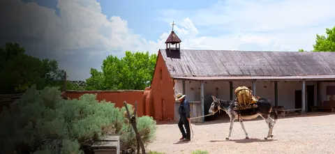 A man leads a pack mule past a historic adobe building with a small bell tower.