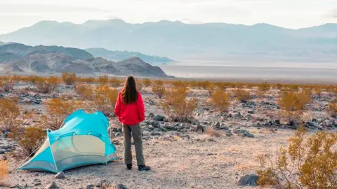 A person standing near a tent in a desert landscape with mountains in the background.
