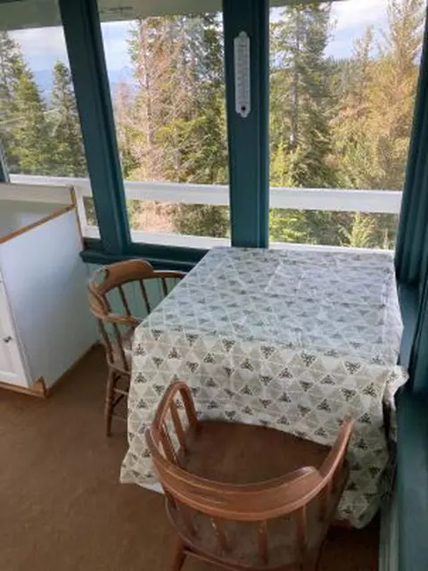 A table and two wooden chairs inside a room with large windows overlooking a forest.