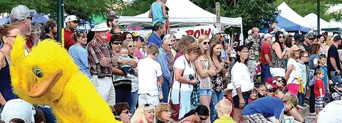 A crowd of people gathered outdoors watching an event, with a large yellow duck mascot in the foreground.