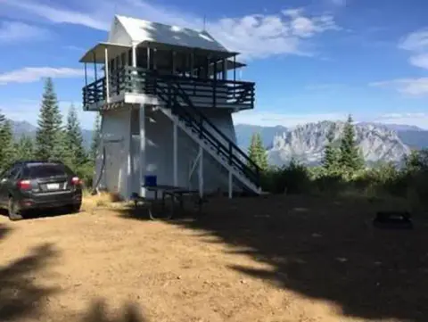 A white lookout tower with dark railings stands on a mountain peak with a car parked nearby.