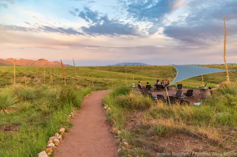 A dirt path leads to a group of people gathered under a shade canopy in a grassy field at sunset.