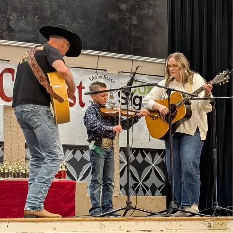 Three musicians perform on a stage with guitars and a fiddle.