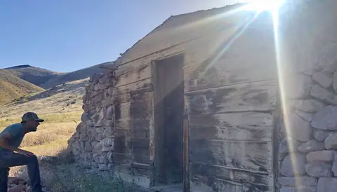 A person stands outside an old wooden and stone building in a dry landscape.